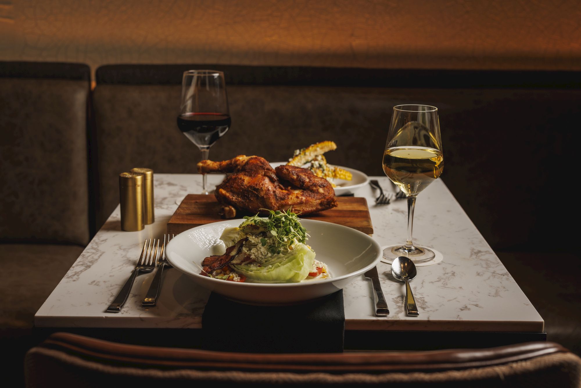 A romantic table for two with wine, a plated dish topped with greens, a roasted bird, and warm lighting in a cozy dining setup.