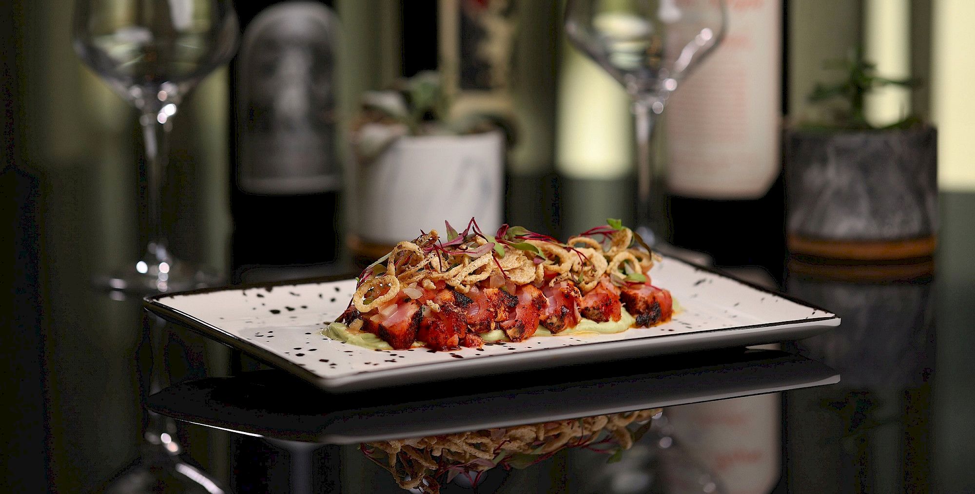 The image shows a plated, gourmet dish with a garnish, set on a reflective surface, surrounded by wine glasses and bottles in the background.