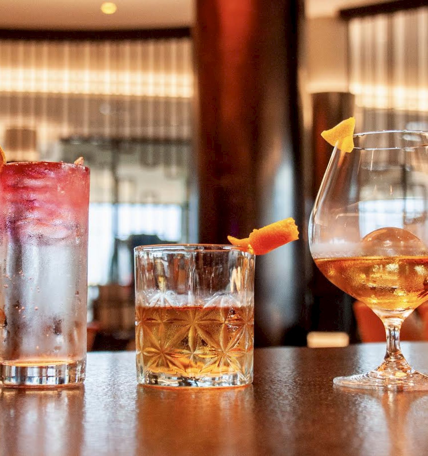 The image shows three different cocktails arranged on a bar counter, each garnished with fruits and seen in a modern interior setting.