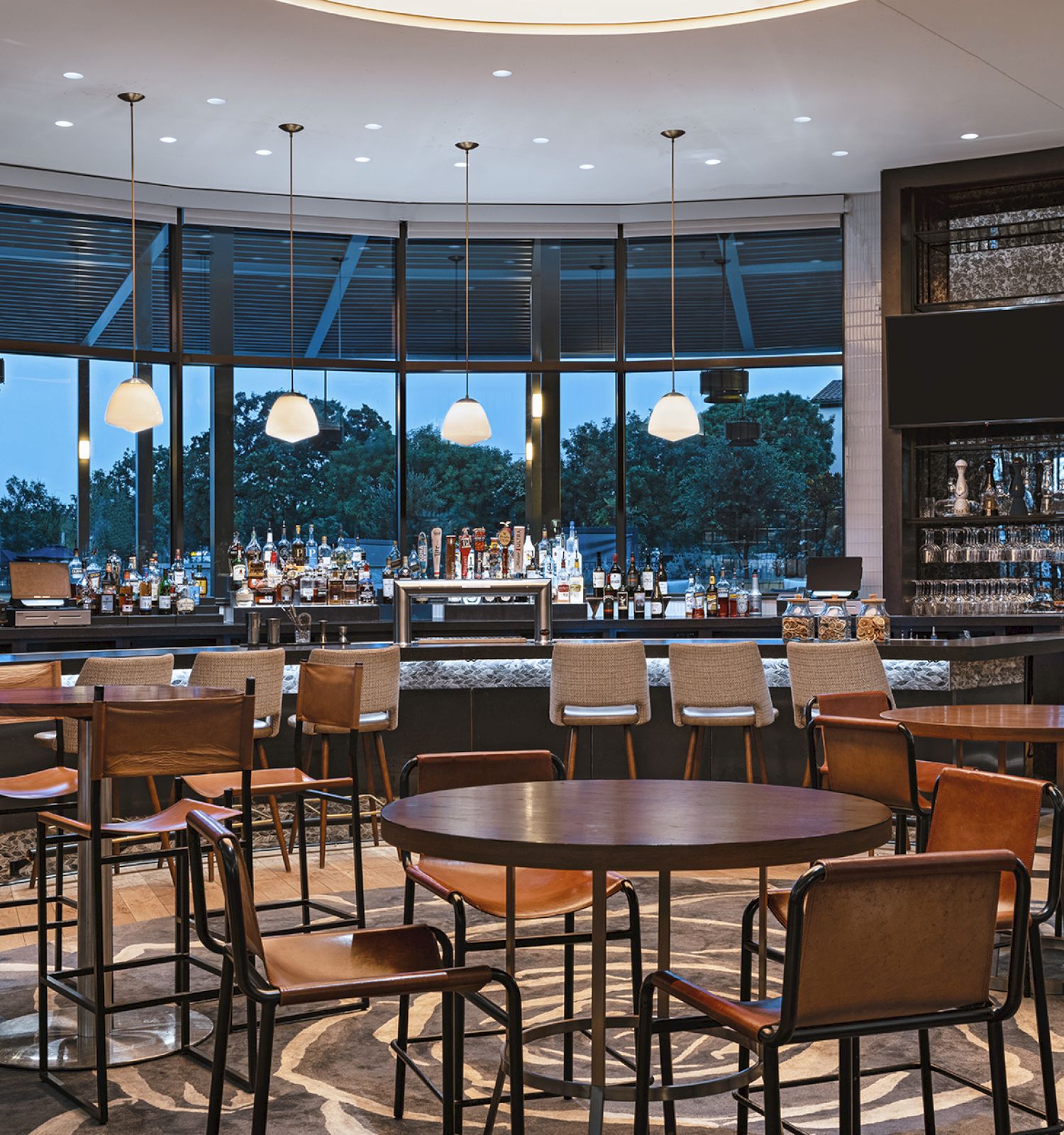 A modern, stylish cafe or bar with wooden tables and leather chairs, a long bar counter, large windows, warm lighting, and a relaxed dining vibe.