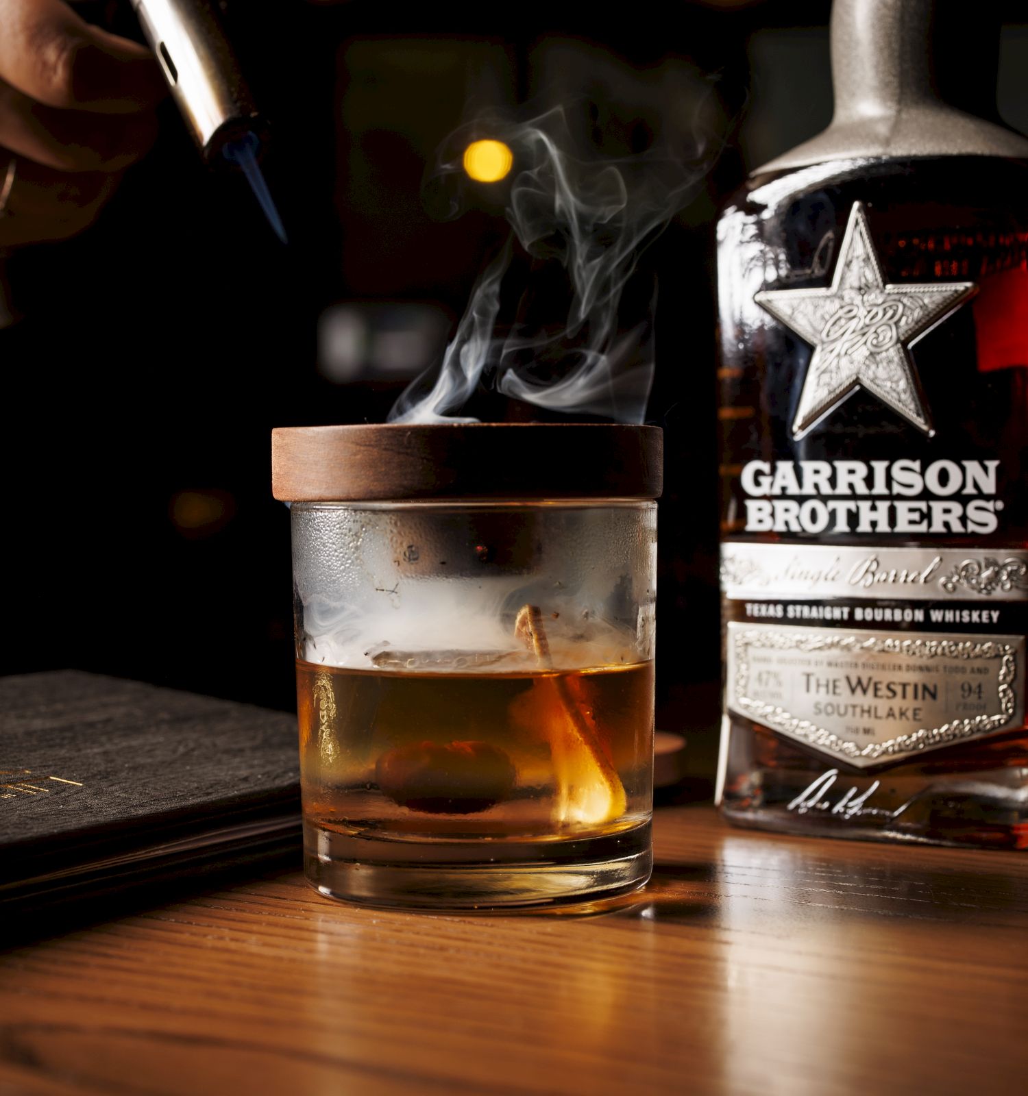 A man pours bourbon into a rocks glass as the drink smokes, with a Garrison Brothers bottle glowing on a bar.