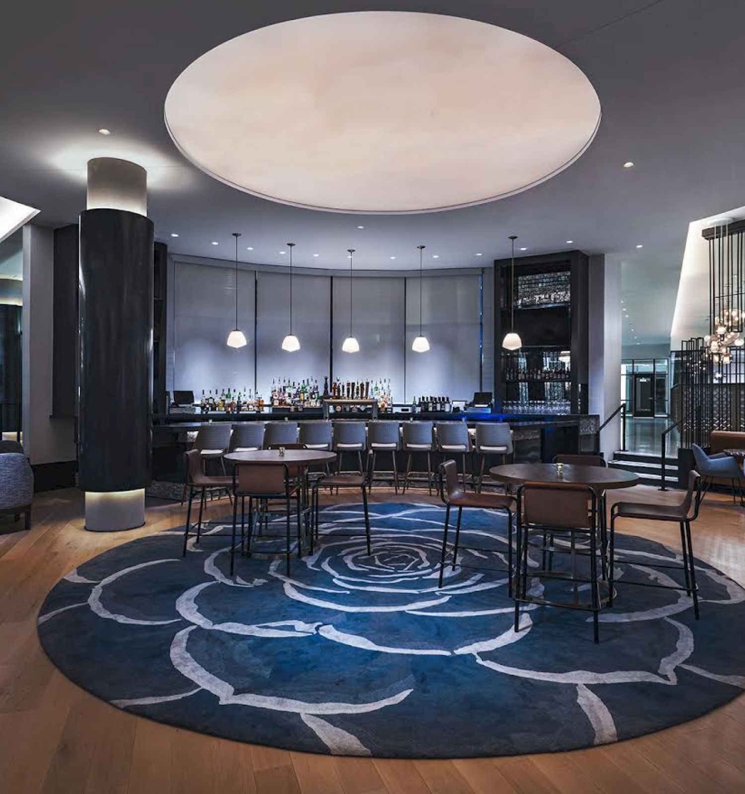 A modern bar with a circular rug, round tables, and pendant lights. The ambiance is sleek with a combination of blue and neutral tones throughout the decor.