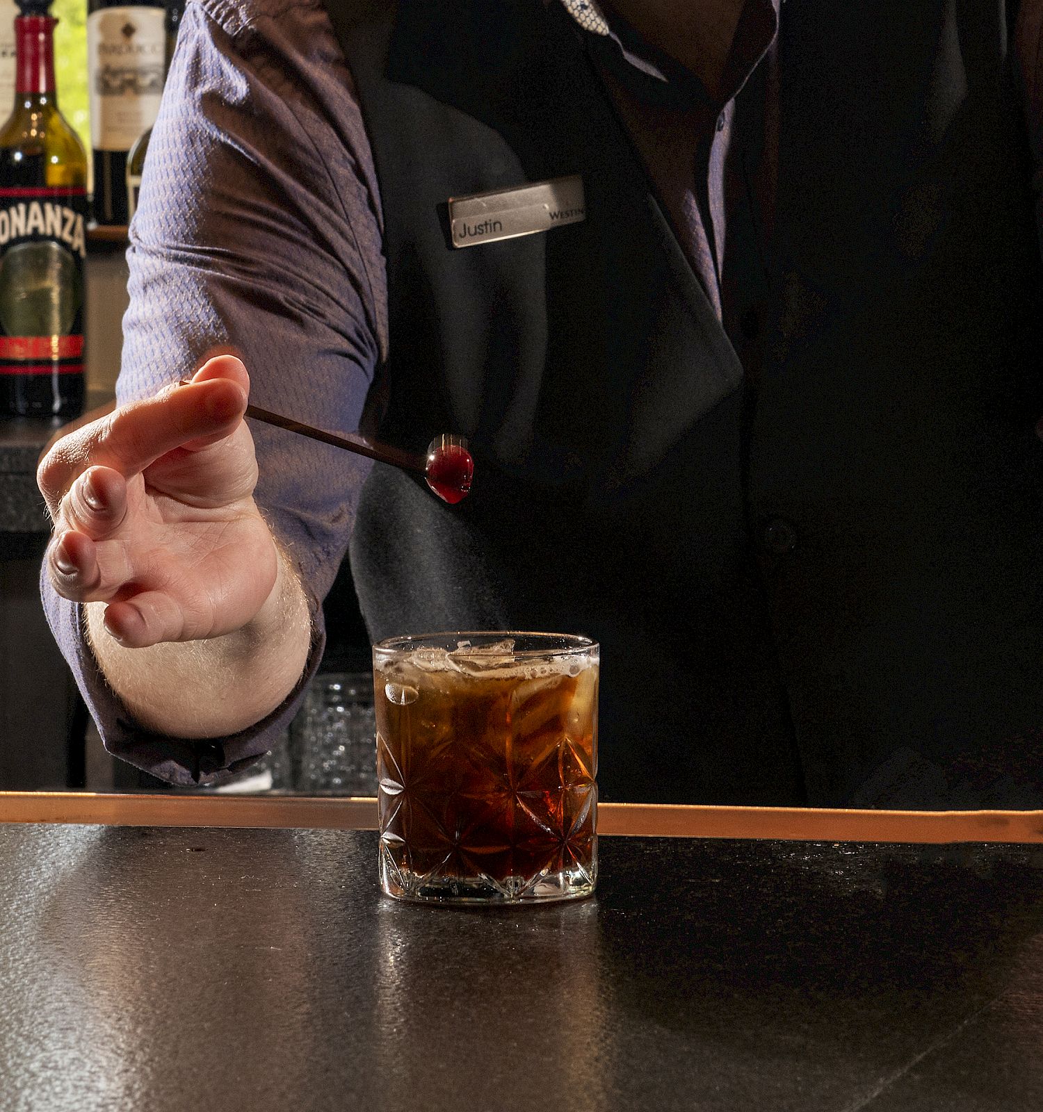 A bartender with a beard reaches for a cocktail glass on the bar, ice cubes clinking, bottles in the background, pointed finger toward the drink.