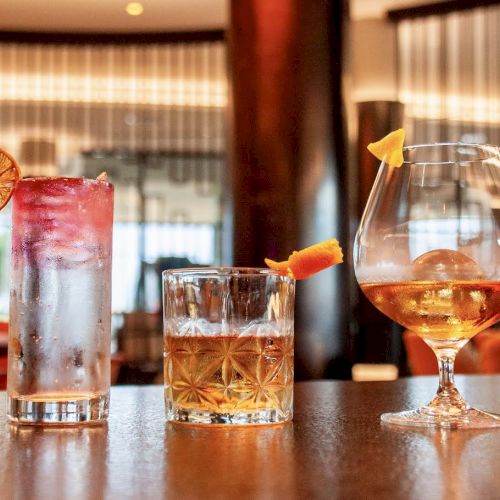 Three different cocktails are lined up on a bar: one with a dried citrus wheel, one with an orange peel, and one in a snifter glass.