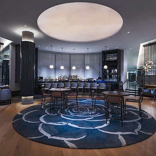 The image shows a modern bar lounge with dark decor, round tables, chairs, a large circular rug, pendant lights, a bar counter, and seating areas.