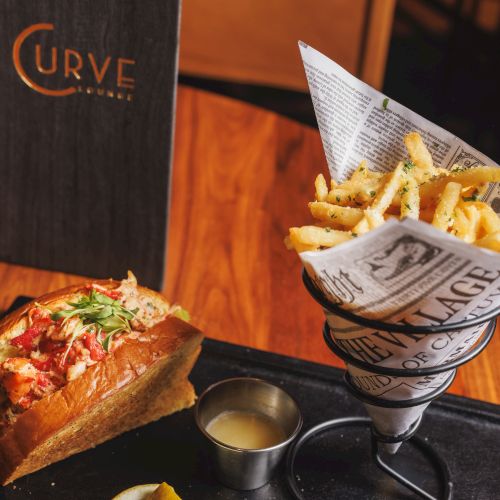 A tray of fries in a paper cone, a slice of cheesy garlic bread, small dipping cups, and a &ldquo;CURVE&rdquo; sign in a restaurant.