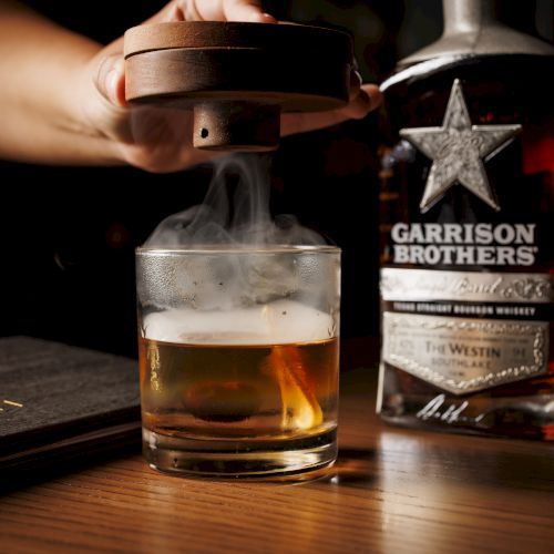 A hand pours whiskey from a &ldquo;Garrison Brothers&rdquo; bottle into an ice-filled glass on a wooden bar, with steam rising.