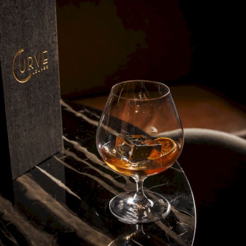 A glass of amber whiskey sits with ice beside a dark &ldquo;CURVE&rdquo; bottle on a textured tabletop.
