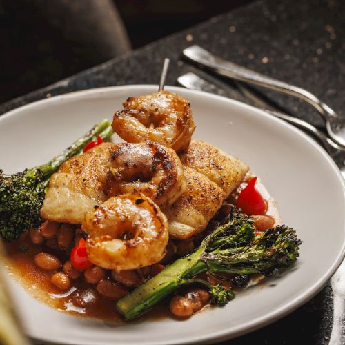 A plated dish of grilled shrimp with sauce, broccoli, carrots, and herbs, served on a dark table with cutlery.