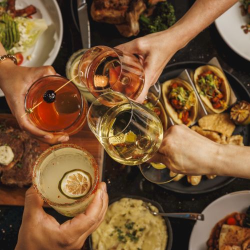People clink glasses of wine/juice over a table of roasted food and garnished bowls, making a celebratory toast.
