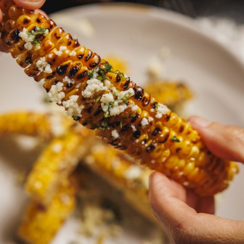Street corn (elote) on the cob, dipped in spicy orange sauce with cotija cheese, chili, and cilantro, being eaten.