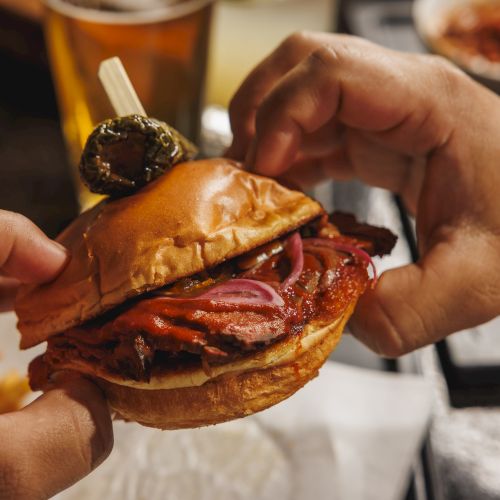 Two hands tearing into a messy burger with melted meat, red onion, and sauce, as a skewer stick is visible.