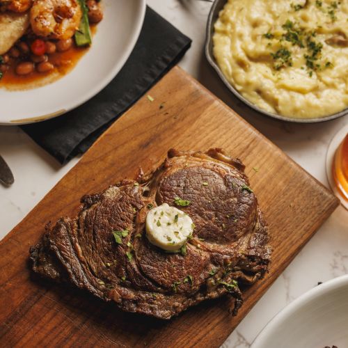 Seared steak with herb butter on a wooden board, alongside creamy mashed potatoes, saucy veggies, and a small cup of sauce.