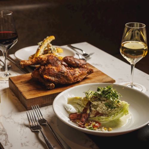 A plated dinner: roasted chicken on a wooden board with roasted garlic, wine glasses, and a side salad with a fork and knife.