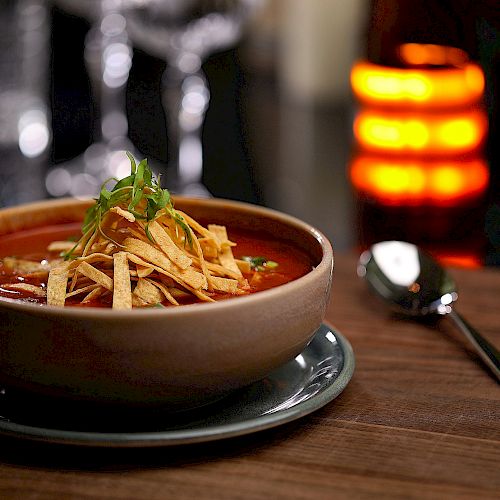 A bowl of soup garnished with tortilla strips and herbs on a wooden table, with a spoon beside it and a blurred background featuring a lit candle.