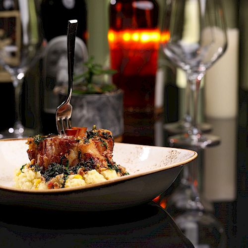 The image shows a plate of food with a fork inserted, surrounded by wine glasses and bottles on a table with a dimly lit, elegant setting.