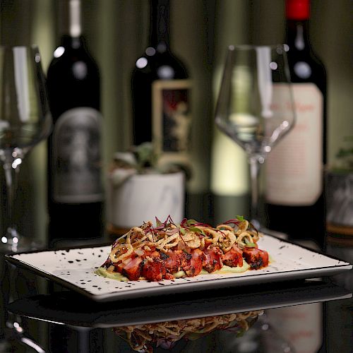 This image shows a plate of food, likely a pasta dish, on a table with two empty wine glasses and several bottles of red wine in the background.