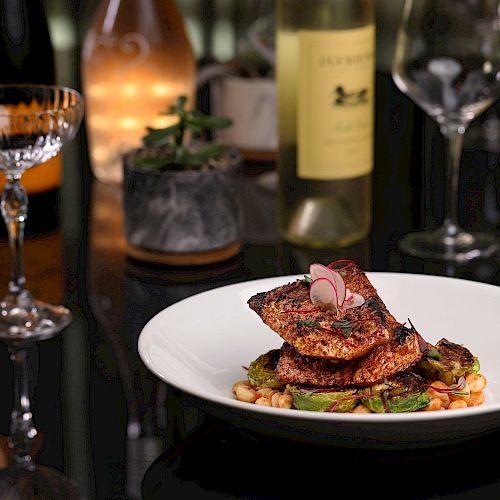 A gourmet dish is served on a white plate, garnished with radishes, with wine glasses, a bottle of white wine, and a lit candle in the background.