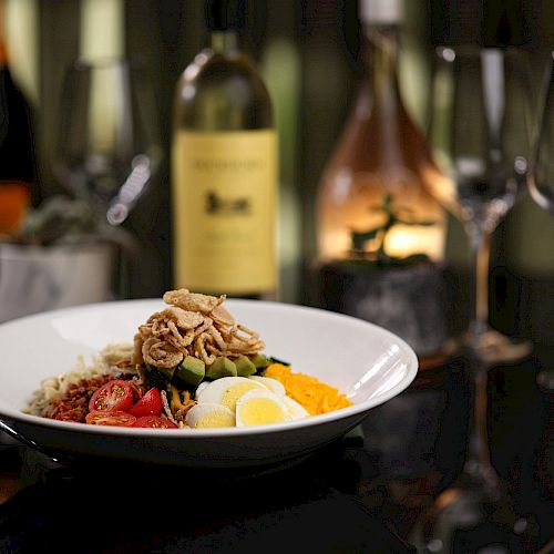 The image features a dish with various vegetables and garnishes in the foreground, with wine bottles and empty wine glasses in the background.