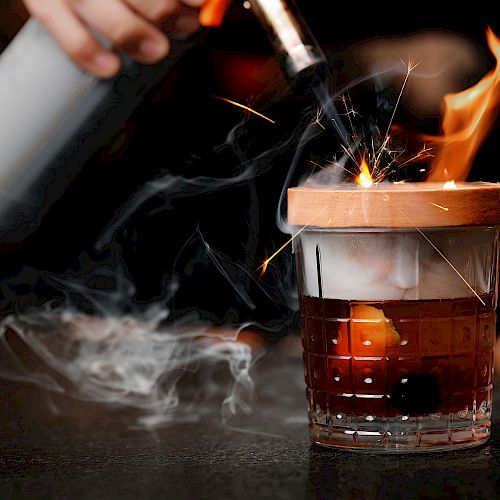 A hand uses a torch to create flames and sparks on a wooden lid over a glass of dark liquid, emitting smoke.