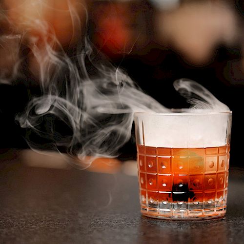 A glass of cocktail with a cherry at the bottom, emitting smoke on a dark surface.