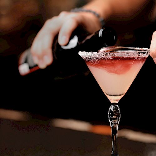 A person is pouring a red liquid into a cocktail glass with a salted rim, in a dimly lit setting.