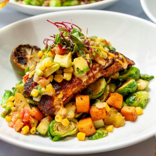 A grilled fish dish topped with a corn and avocado salsa, served with mixed vegetables and a charred lemon on the side.