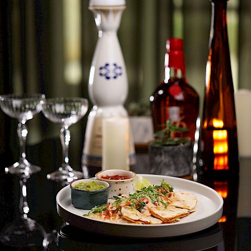 The image shows a sophisticated table setting with drinks, glasses, a candle, and a plate of quesadillas accompanied by two dips.