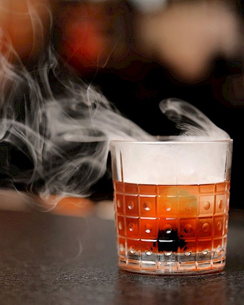 A glass containing a beverage, possibly a cocktail, with smoke or vapor rising from it, placed on a dark surface in a blurred background setting.