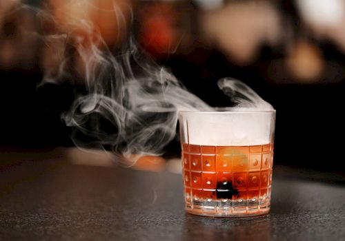 A glass containing a beverage, possibly a cocktail, with smoke or vapor rising from it, placed on a dark surface in a blurred background setting.