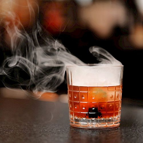 A glass containing a beverage, possibly a cocktail, with smoke or vapor rising from it, placed on a dark surface in a blurred background setting.