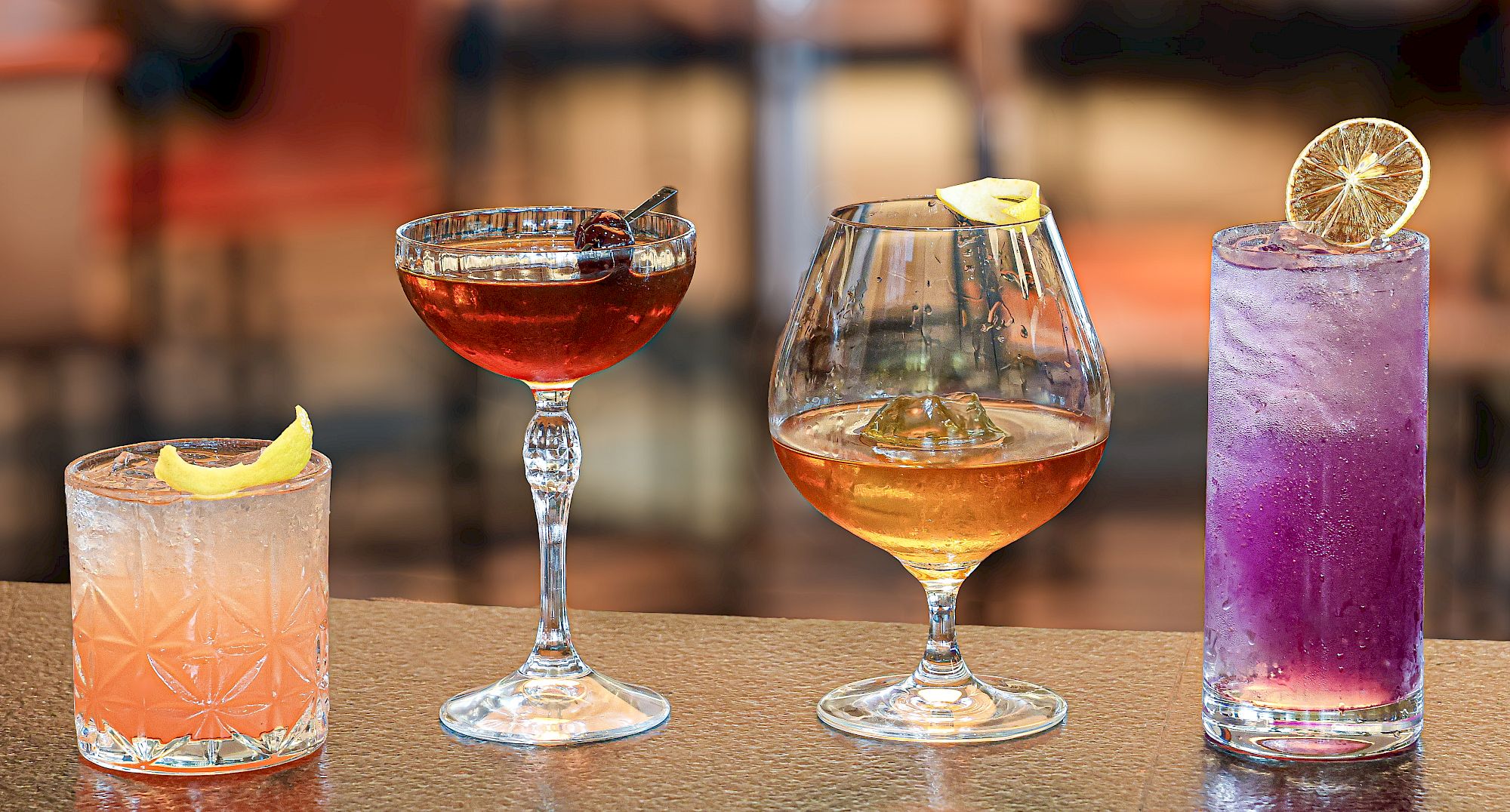 A row of four cocktails on a bar: a pink citrus drink, a dark red cocktail in a coupe, a golden whiskey-style glass, and a tall purple lavender drink with a lemon slice on top.