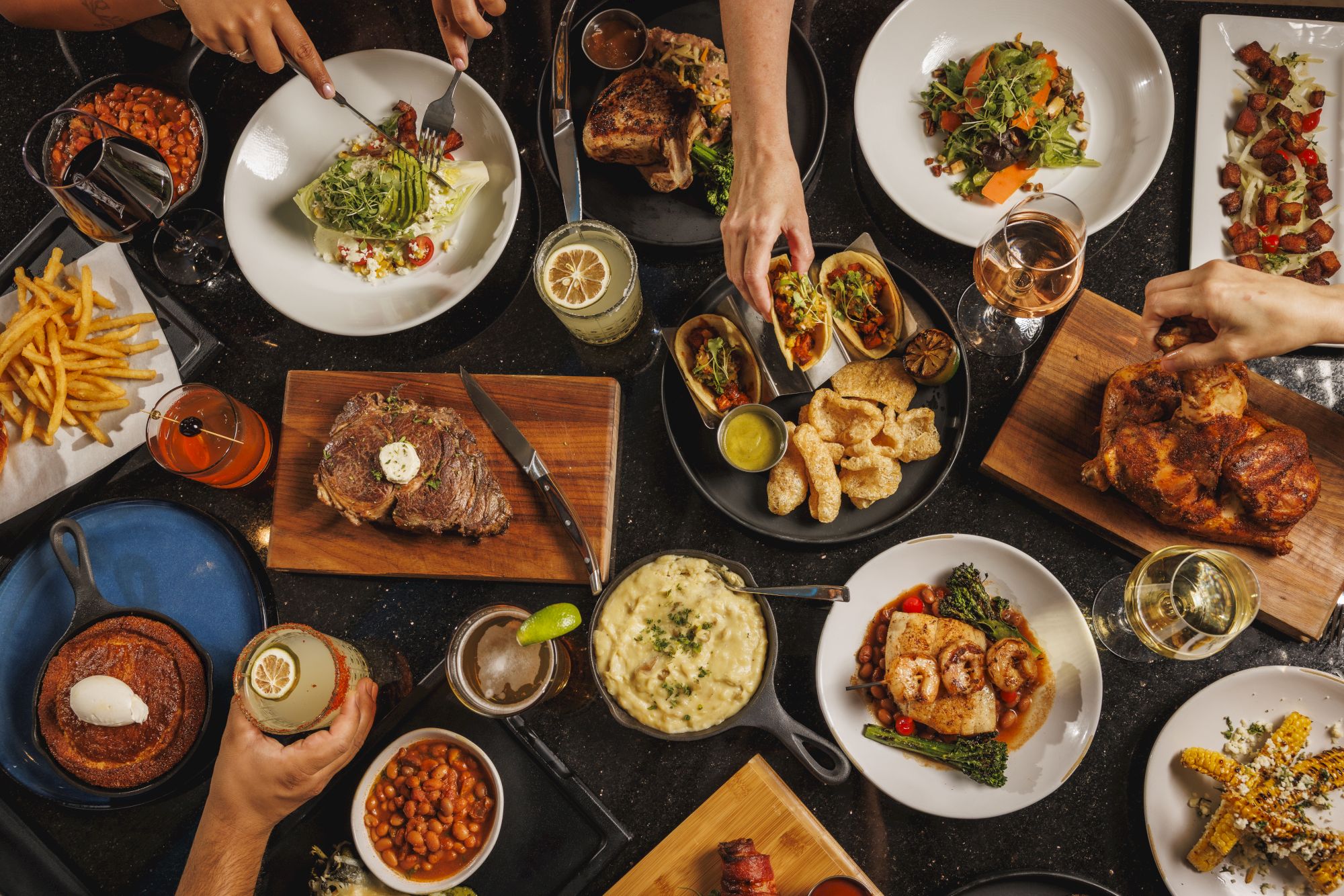 A table full of assorted dishes and hands reaching in: steak, tacos, fries, salads, roasted chicken, dips, sauces, and drinks on a busy, shared meal.