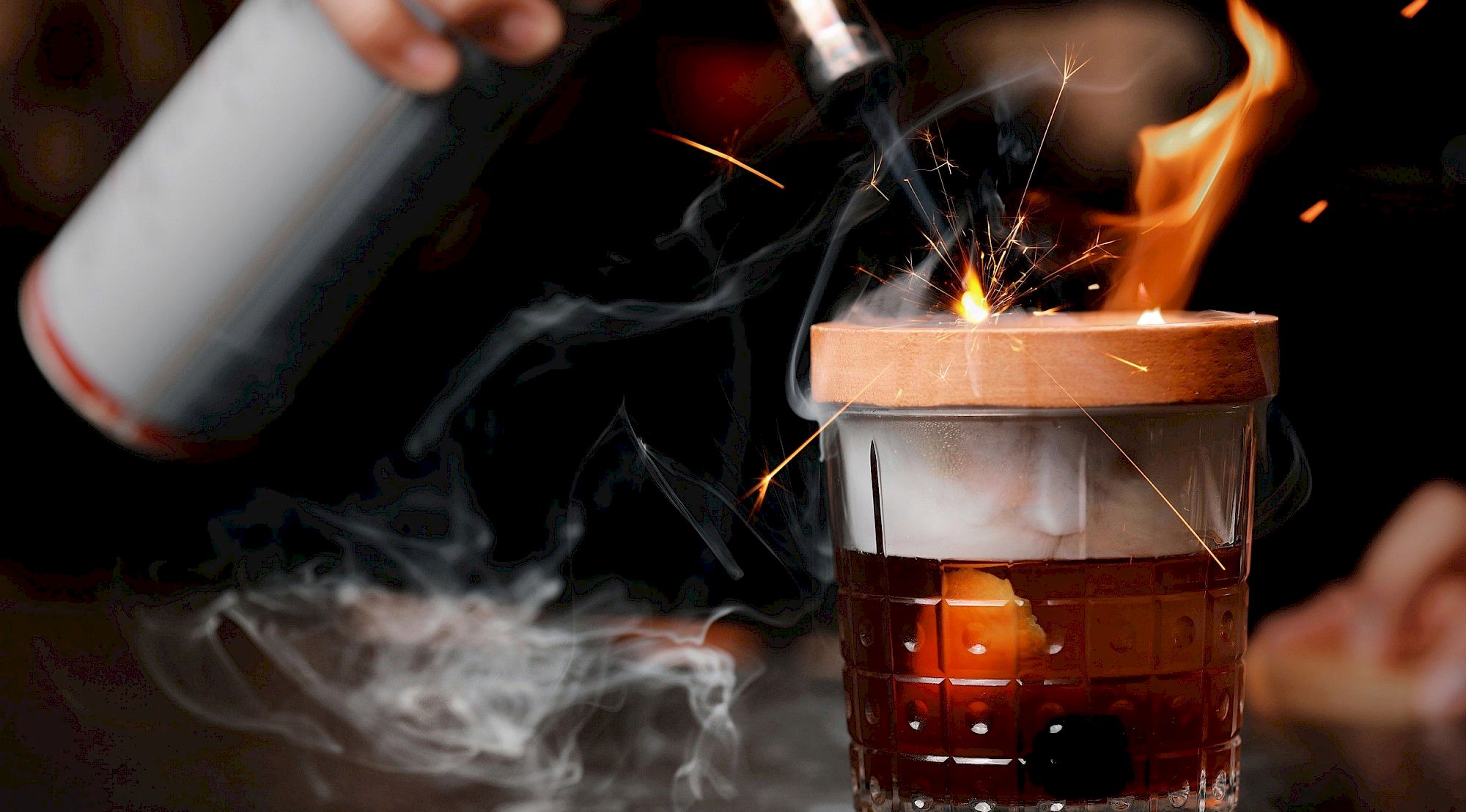 A person is igniting a cocktail with a torch, causing flames and sparks, while the drink is adorned with a wooden coaster releasing smoke.