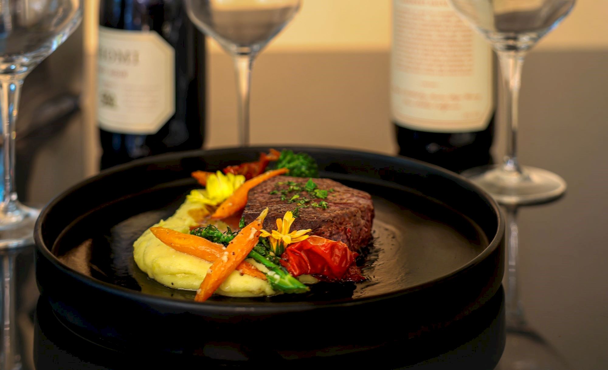 A gourmet dish with steak, mashed potatoes, and vegetables on a black plate, surrounded by wine bottles and empty glasses.