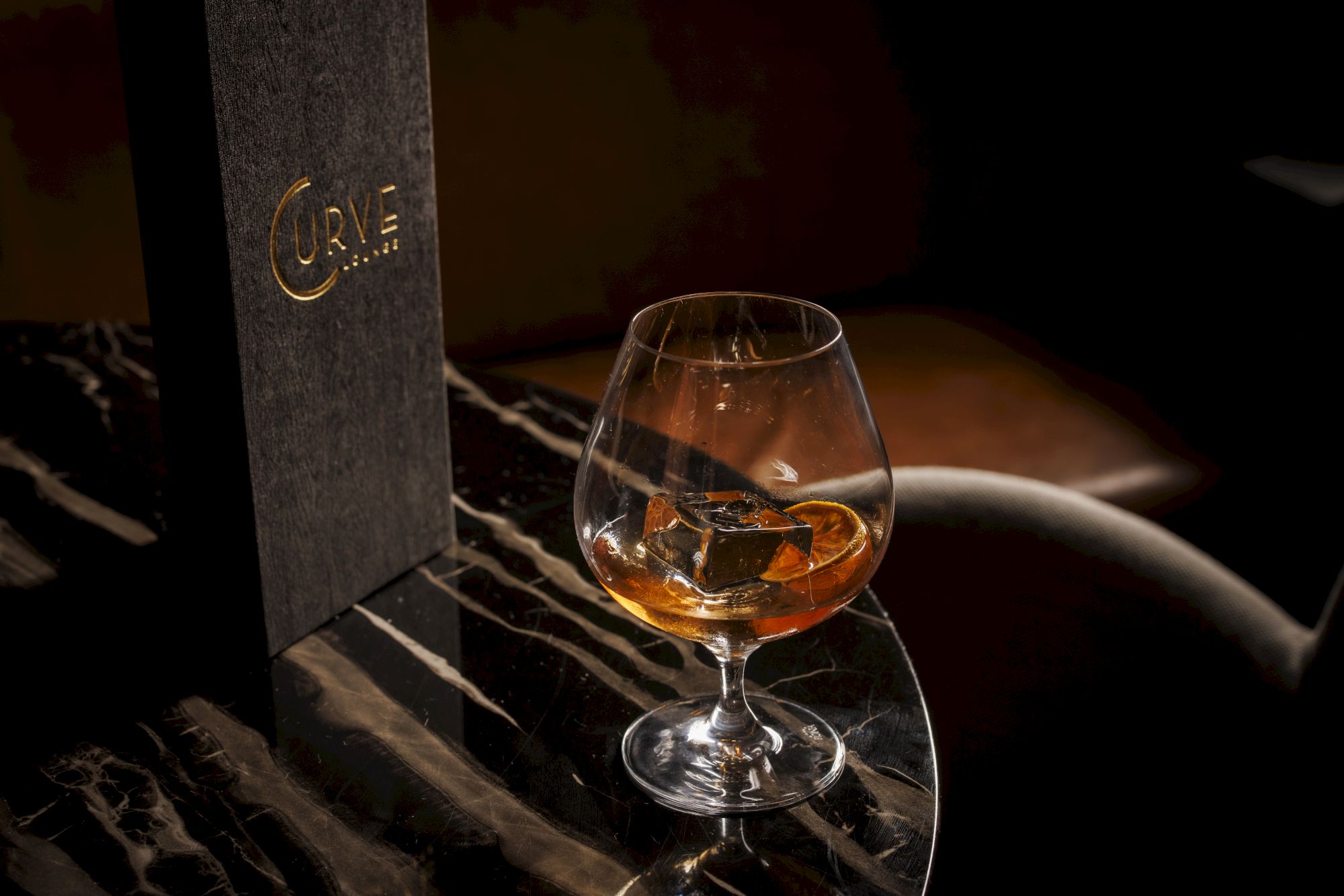 A snifter of amber spirit on a marble table beside a dark menu, in a moody bar setting.