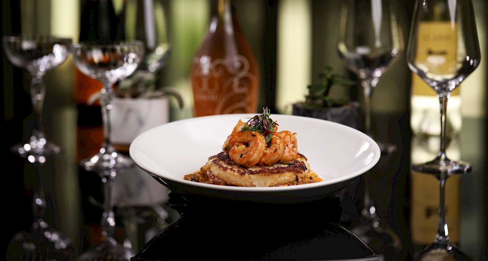 A restaurant table with a white bowl filled with a gourmet shrimp dish, surrounded by wine glasses and a bottle in the background.