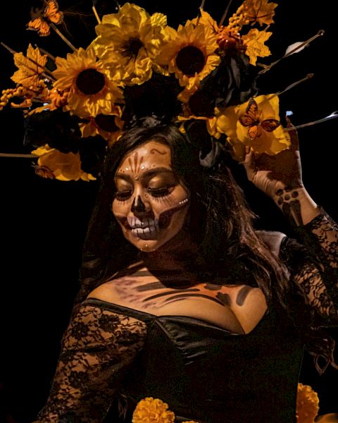 A person wearing a floral headpiece and skull face paint, dressed in a black lace outfit, posed dramatically against a dark background.