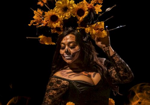 A person wearing a floral headpiece and skull face paint, dressed in a black lace outfit, posed dramatically against a dark background.