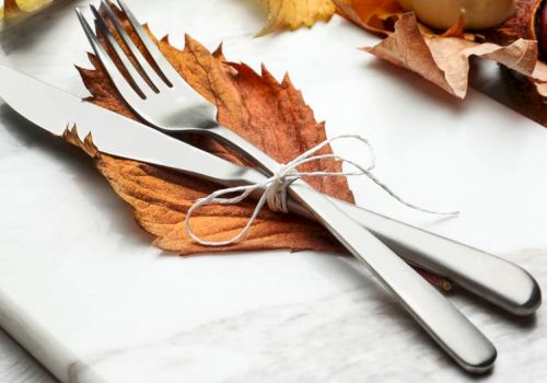 A knife and fork tied with string rest on autumn leaves, surrounded by more leaves and a small pumpkin on a marble surface.