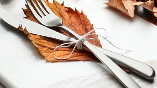 A knife and fork tied with string rest on autumn leaves, surrounded by more leaves and a small pumpkin on a marble surface.