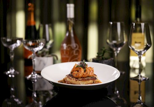 A plate of gourmet food is in focus, with shrimp on toast, surrounded by wine glasses and bottles in the background.