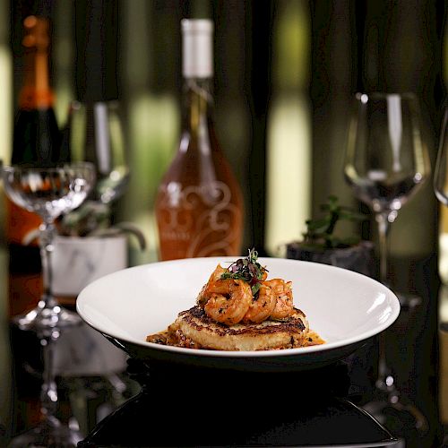 A plate of gourmet food is in focus, with shrimp on toast, surrounded by wine glasses and bottles in the background.