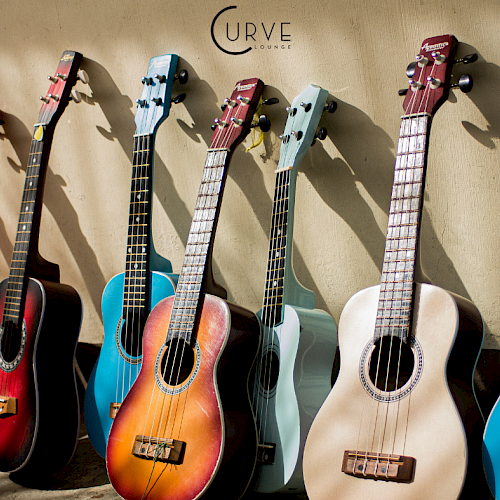 The image shows a row of colorful guitars and ukuleles leaning against a wall, with the word "CURVE" and a logo above them.