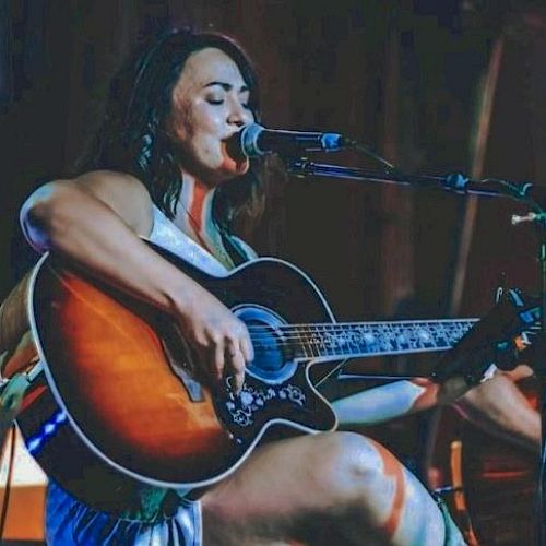 A person is seated while playing an acoustic guitar and singing into a microphone on stage, with a dark background.