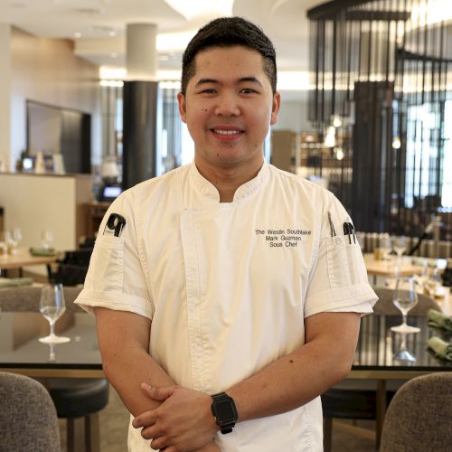 A chef in a white uniform is standing in a restaurant, smiling, with crossed arms.