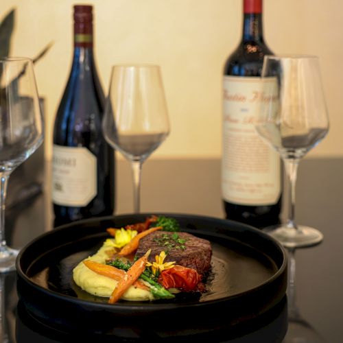 A gourmet dish with steak, mashed potatoes, and vegetables is paired with two wine bottles and empty glasses on a table.