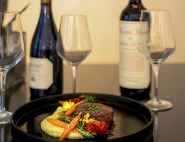A gourmet dish with steak, mashed potatoes, and vegetables is paired with two wine bottles and empty glasses on a table.