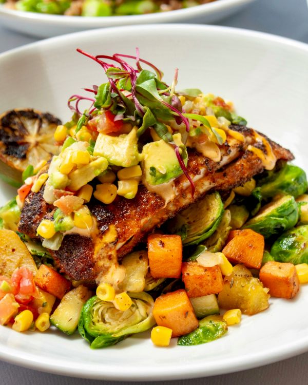 A plated dish featuring seared fish topped with a vegetable salsa, served over roasted vegetables including Brussels sprouts and diced squash.