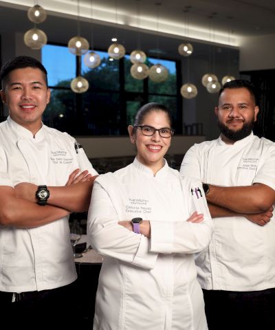 Three chefs wearing white uniforms stand with arms crossed, smiling in a well-lit restaurant setting.